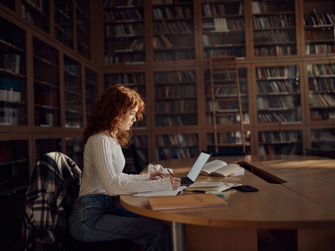 Teenage female student taking notes while using a computer in library.
